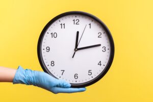 Profile side view closeup of human hand in blue surgical gloves holding analog clock.